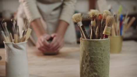 una alfarera amasa arcilla en la mesa en un estudio de arte - pinceles y otras herramientas en primer plano