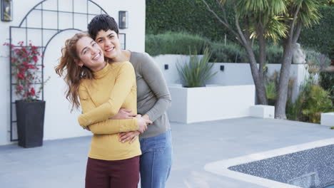 una feliz pareja de lesbianas caucásicas de pie, abrazándose y sonriendo frente a la casa