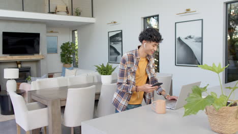 using smartphone and laptop, teenage boy working from home in modern living room
