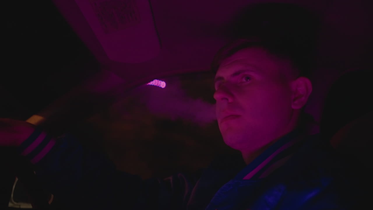 Interior nocturno de un coche con un conductor exhalando vapor en primer plano de perfil, luz púrpura bañando la cara y nube llenando la cabina. Detalle de chaqueta de cuero, vislumbres del espejo retrovisor, crucero lento por la ciudad con reflejos.