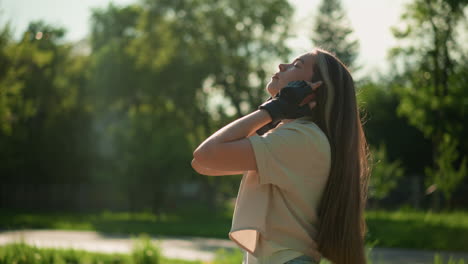vista lateral de una mujer ciclista ajustando el cabello y la parte superior, de pie con la cabeza inclinada hacia atrás bajo la cálida luz del sol, el fondo presenta vegetación y árboles suavemente enfocados