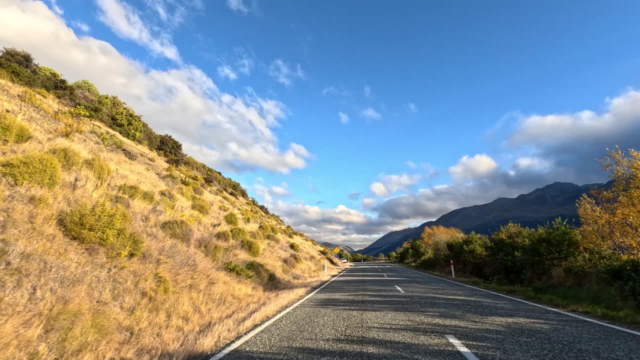 Car travels along sunlit mountain road, passing golden hills and greenery under blue sky