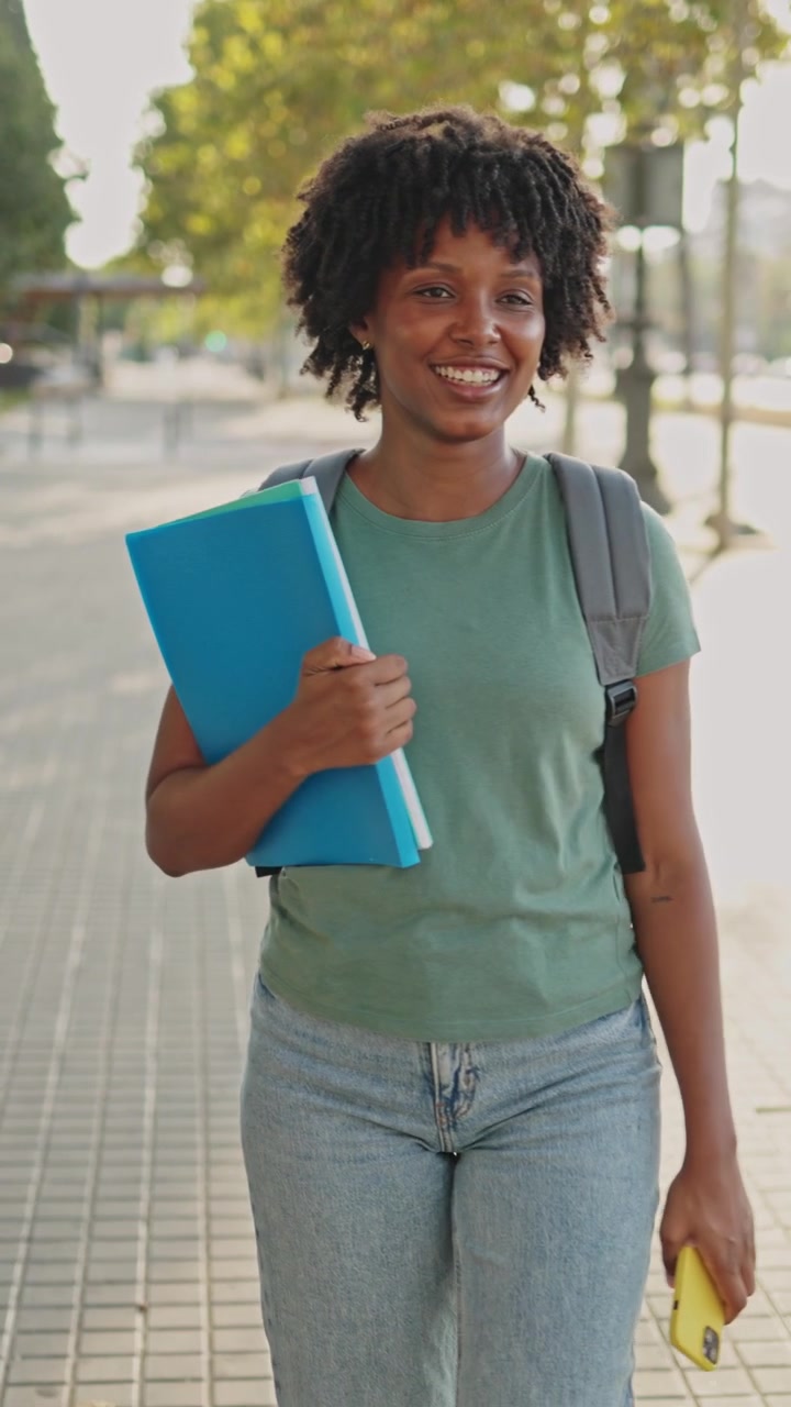 Studentessa afroamericana sorridente cammina nel campus con cartelle e telefono