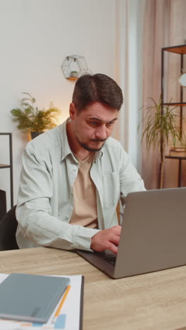 caucasian young man freelancer opens laptop and working on project sitting at home office table