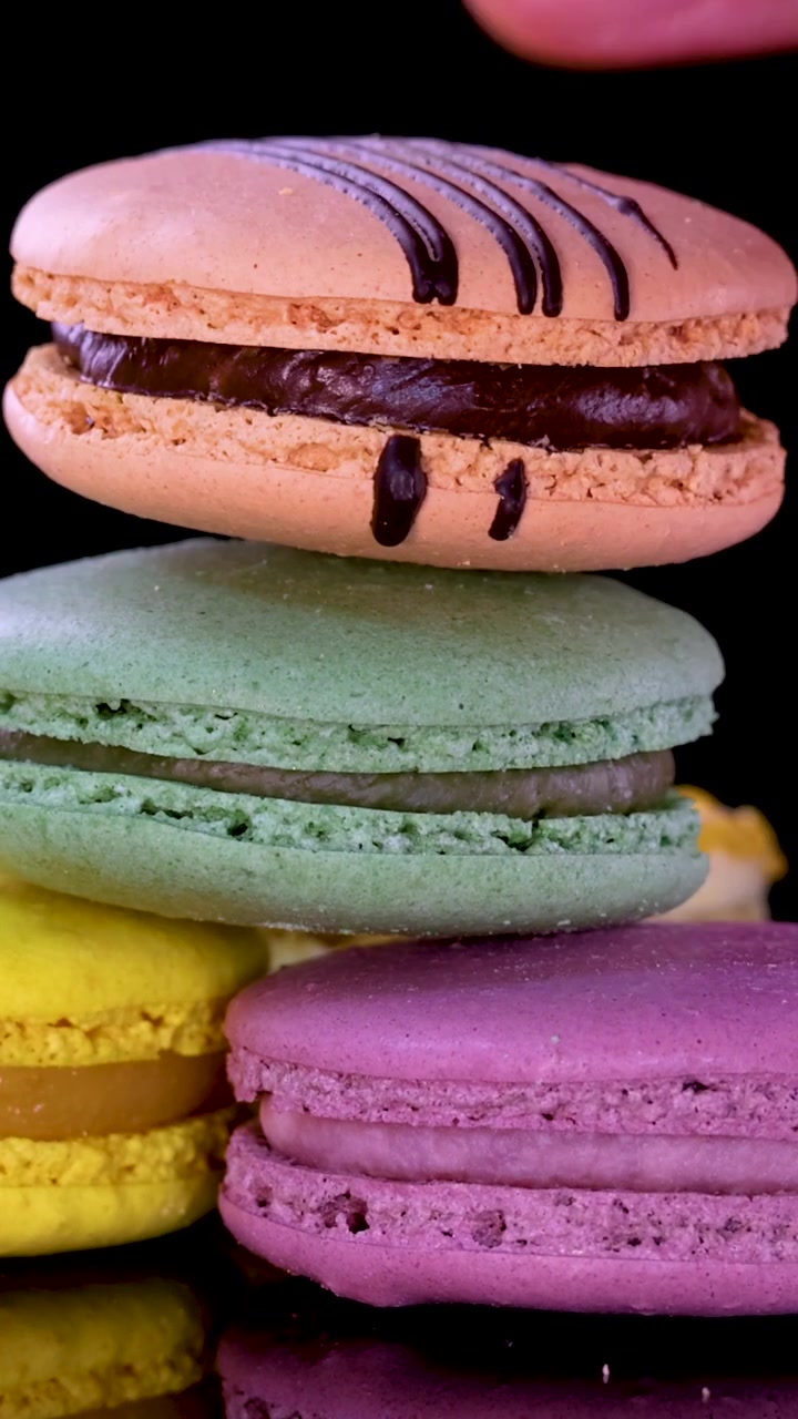 A hand carefully stacks vibrant green, purple, and chocolate drizzled macarons against a dark background.