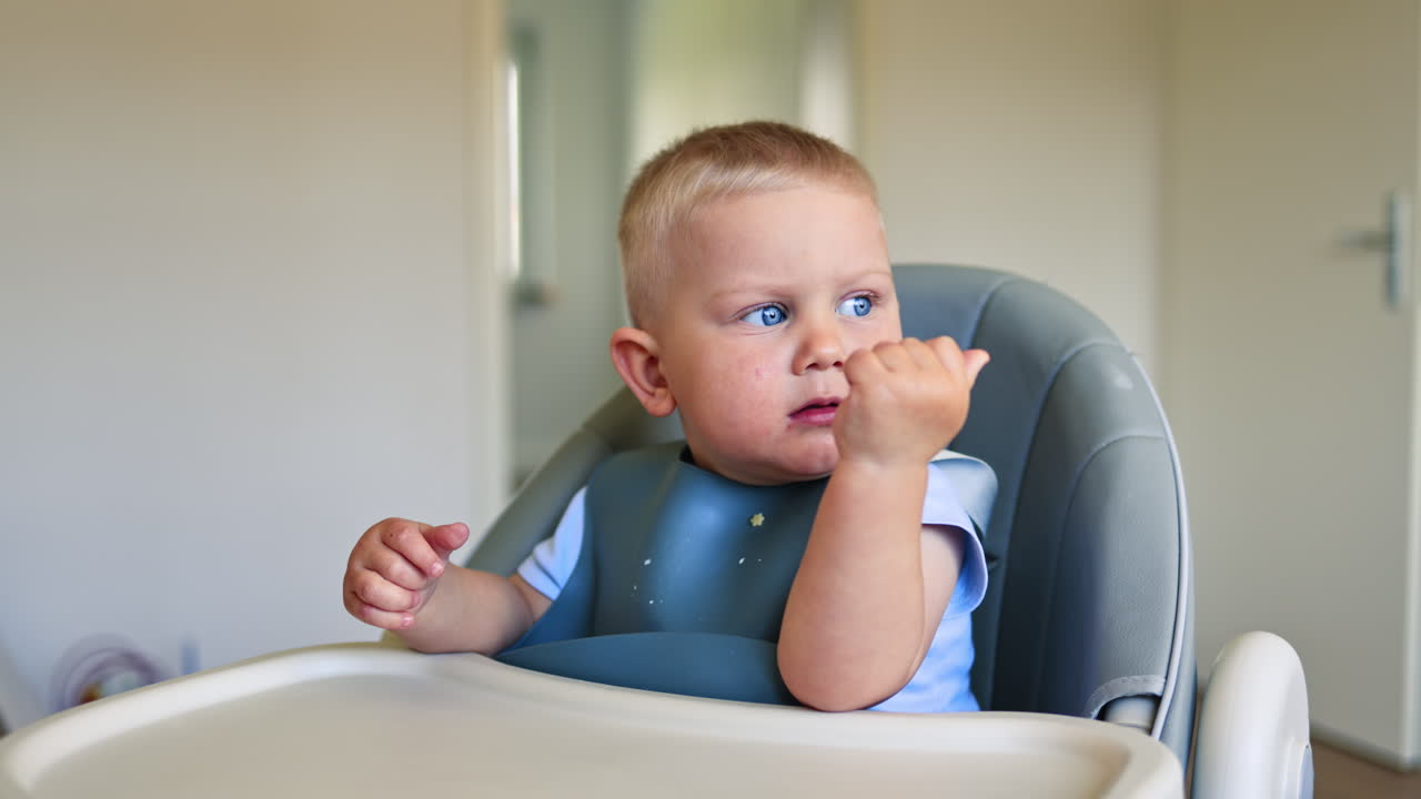 Adorable blond boy with silicone utensils sits on a high chair for feeding. Joyful child drinks deliciously and smiles. Child development. Children's furniture for feeding