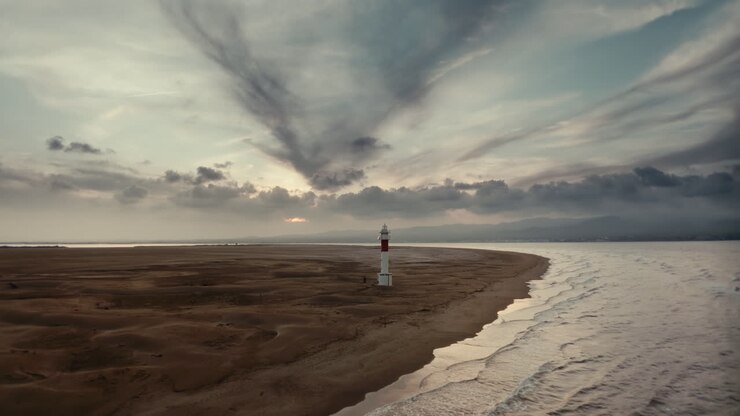 Smukke 4K droneoptagelser af et ensomt fyrtårn på en bred sandstrand ved solnedgang, med dramatiske skyformationer over hovedet og blide bølger, der ruller ind, hvilket skaber en fredelig filmisk kystscene.