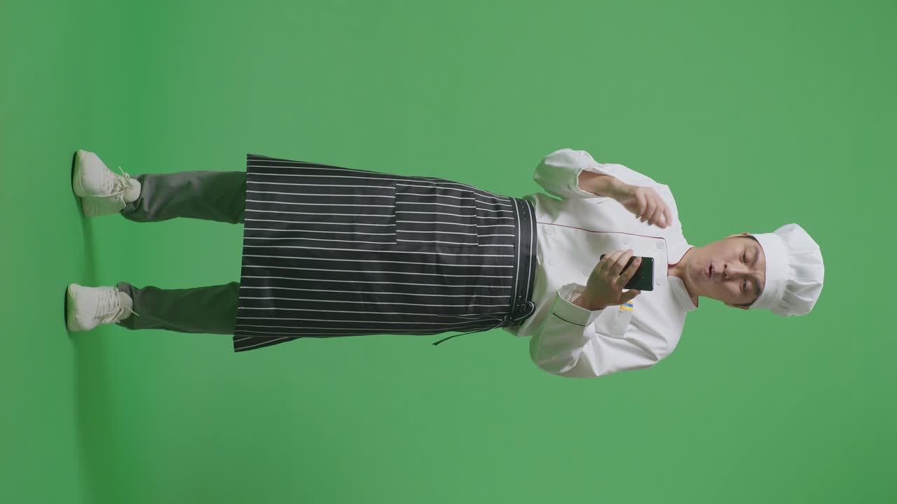 Full Body Of Asian Man Chef Looking At Smartphone, Smiling, And Saying Wow While Standing In The Green Screen Background Studio