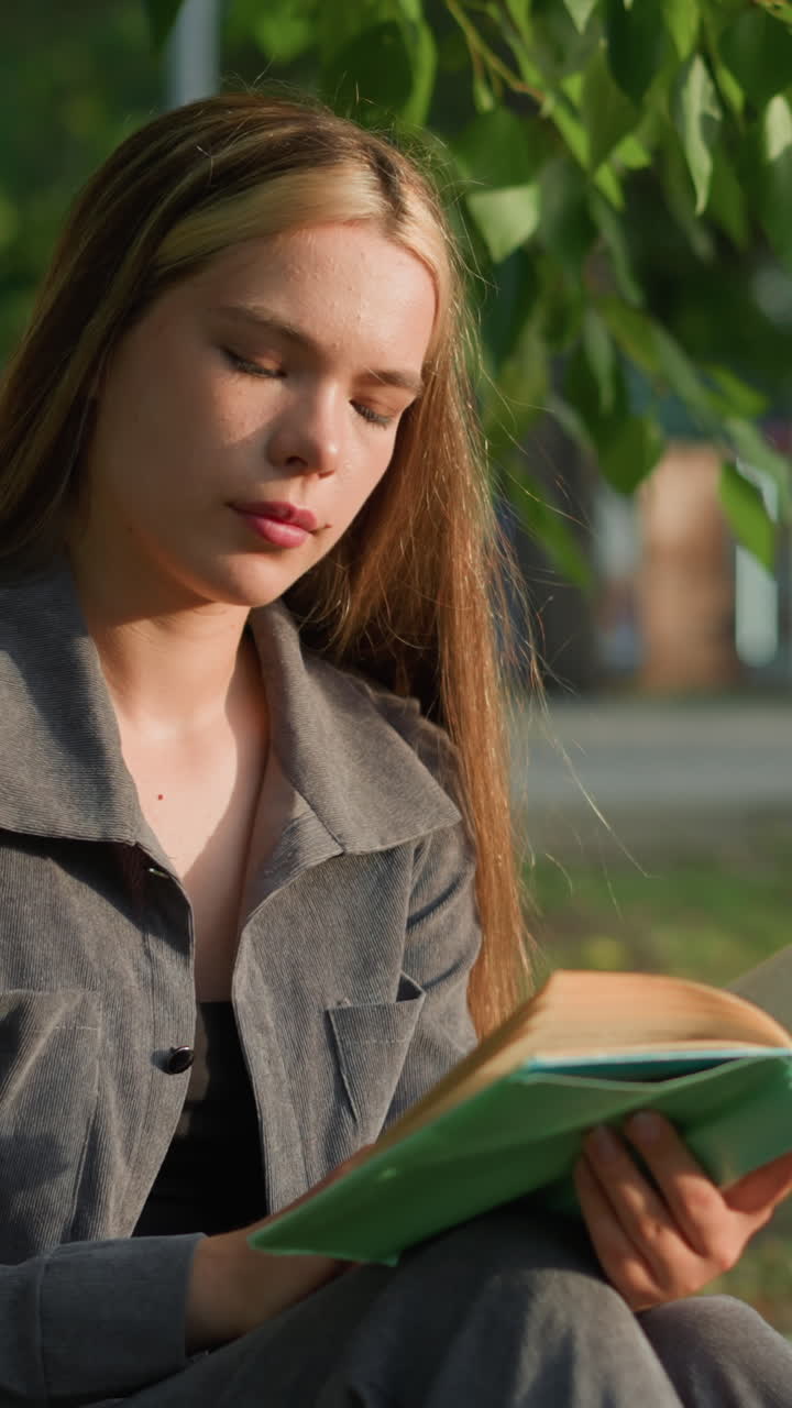 dama con ropa gris sentada al aire libre leyendo, mirando pensativa, el fondo presenta vegetación y edificio borroso, con hojas por encima balanceándose suavemente en el viento