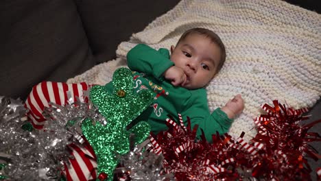 adorable bebé asiático de 2 meses acostado en una manta con un traje navideño festivo chupando la mano izquierda con oropel rojo y plateado a su lado