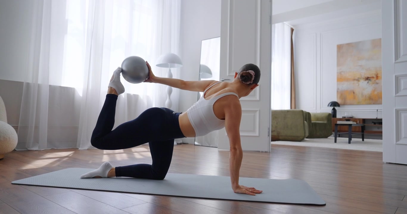 Giovane donna che pratica Pilates con palla piccola su tappetino da yoga a casa