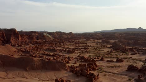 Gorgeous-dolly-out-aerial-drone-shot-of-the-beautiful-Goblin-Valley-Utah-State-Park-with-small-strange-mushroom-rock-formations-below-and-large-red-and-white-Butte's-in-the-background