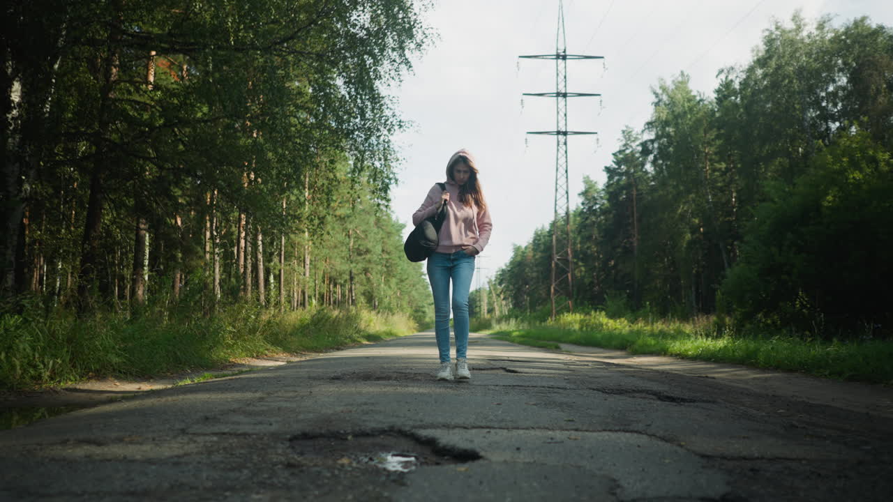 Young lady walking alone on cracked tarred road lined with tall trees and overhead power lines under daylight, carrying black bag, surrounded by serene greenery