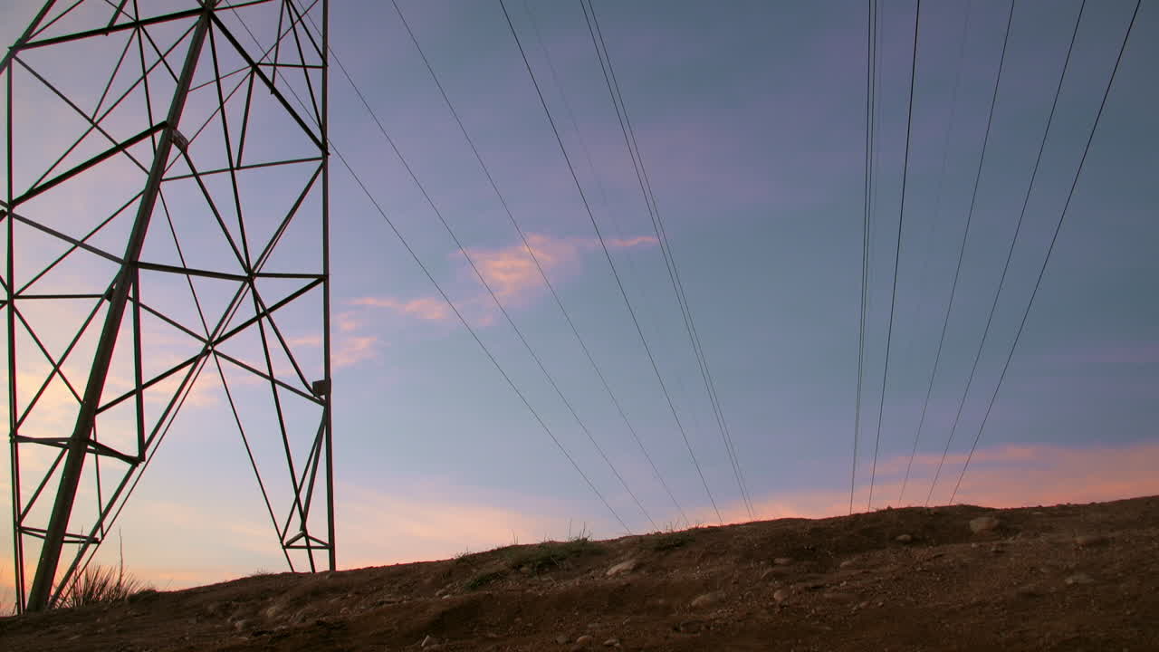 A male mountain biker, performs a wheelie trick, gliding underneath a power line tower, as the low sun silhouettes his form