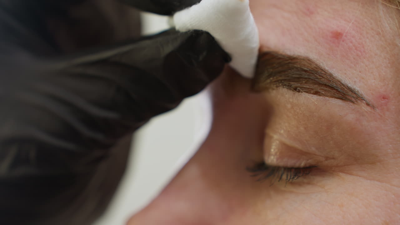 Close up of beautician wearing black glove gently cleaning client's eyebrow with white cotton pad during beauty treatment, showcasing delicate care and precision in personal grooming process