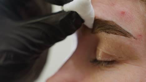 close up of beautician wearing black glove gently cleaning client's eyebrow with white cotton pad during beauty treatment, showcasing delicate care and precision in personal grooming process