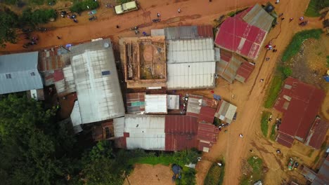Top-down drone descent reveals dense cluster of corrugated iron-roofed structures forming a busy trading hub on a dusty road in rural Uganda, surrounded by vegetation and scattered housing