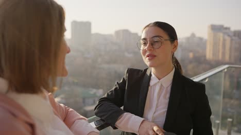 Over-the-shoulder-a-confident-brunette-businesswoman-girl-in-round-glasses-and-a-black-uniform-communicates-with-her-colleague-while-standing-on-the-terrace-in-a-modern-office-overlooking-the-city-sunny-weather