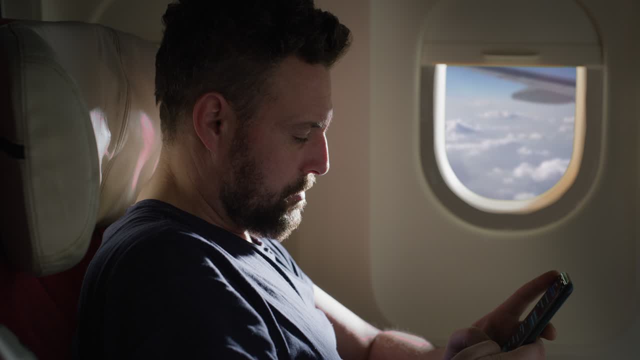 una toma cinematográfica de un joven que usa un teléfono inteligente para enviar mensajes y aplicaciones tecnológicas mediante el uso de conexión inalámbrica en aviones mientras viaja con comodidad durante un vuelo internacional a un viaje de vacaciones.