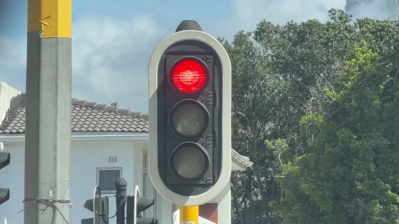 Close de um semáforo mostrando a cor vermelha, montado em um poste com uma casa suburbana e árvores verdes exuberantes ao fundo com foco suave. Iluminação clara de dia