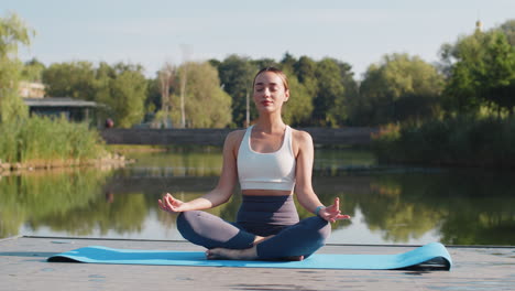 caucasian young woman sitting on mat in lotus position relaxing near pond in park on summer day