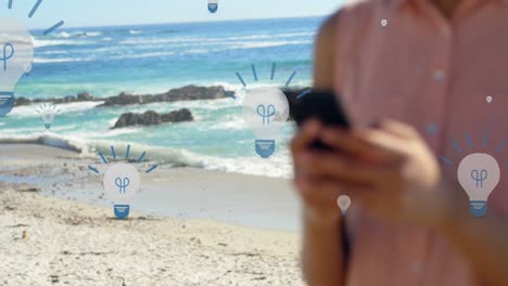 animación de iconos de bombillas sobre la sección media de una mujer biracial que usa un teléfono inteligente en la playa