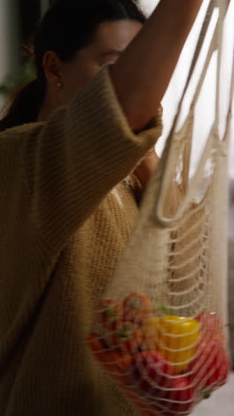 vertical video of woman talking on mobile phone getting home from food shopping unpacking bag of fresh vegetables onto counter in kitchen