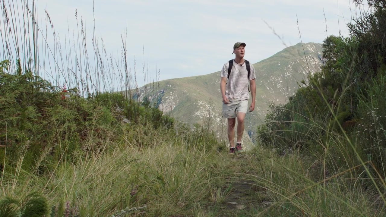 Escursionista solitario che indossa un cappellino e uno zaino avanza lungo un sentiero stretto attraverso erba alta, con montagne verdi ondulate visibili sullo sfondo. Ampia vista statica a livello degli occhi.