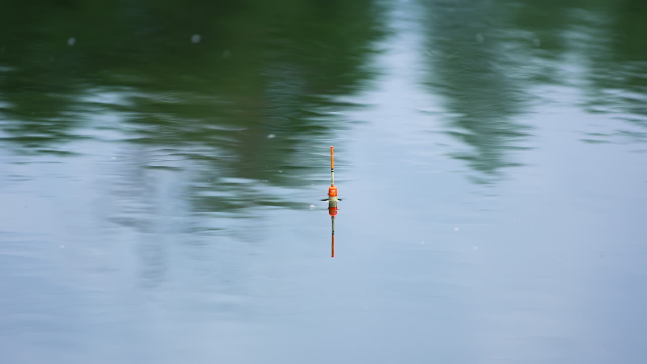 White and orange fishing bobber floating in the river. Unmoving float on still water on cloudy day.
