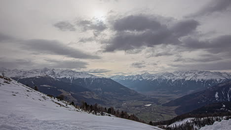 Tiro-De-ángulo-Alto-Desde-La-Cima-De-La-Montaña-Cubierta-De-Nieve-De-Un-Valle-Cubierto-De-Vegetación-Verde-En-Un-Día-Nublado-En-Timelapse