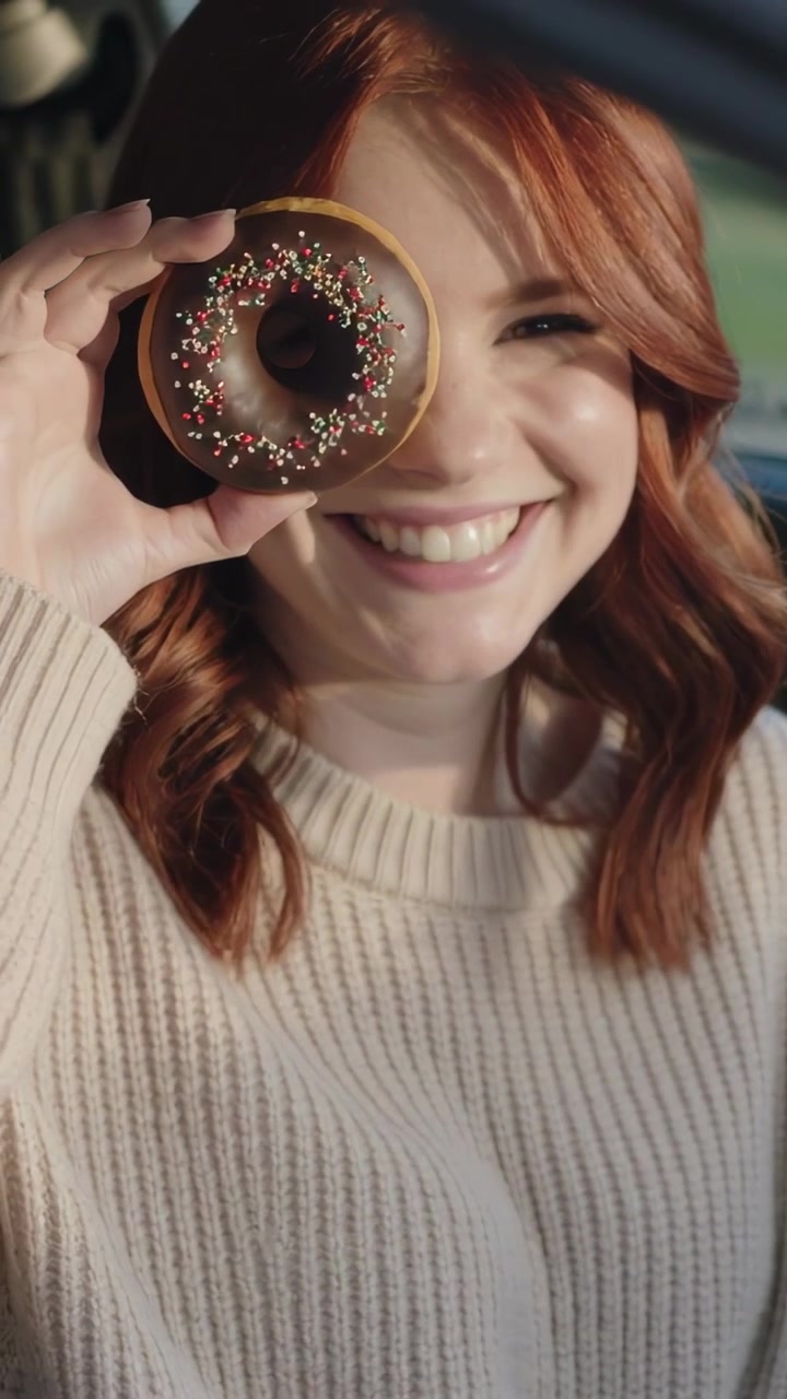 Вертикальное видео: Рыжеволосая женщина в кремовом свитере играет с пончиком, смеется, затем кусает его в машине.