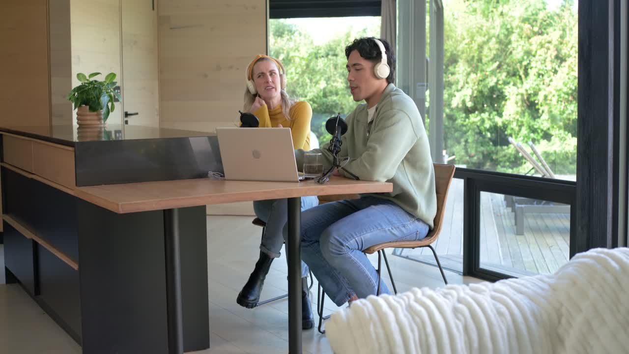 Sunlight streaming sparking cohosts adjusting mic cradling kitten and chatting at laptop in kitchen