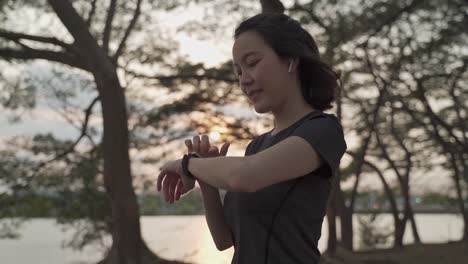 asian athlete woman using sport smart watch green nature park in evening sunset. female in sportswear touching screen application tracking activity and music. enjoy personal record data performance.