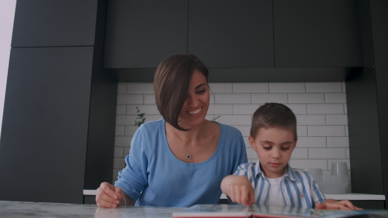 una hermosa madre y su hijo sentados en una mesa en una cocina luminosa leyendo un libro y mirando imágenes metiendo un dedo en un libro y hojeando las páginas
