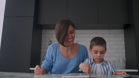 Young-beautiful-mother-and-son-sitting-at-a-table-in-a-bright-kitchen-reading-a-book-and-looking-at-pictures-poking-a-finger-into-a-book-and-leafing-through-the-pages