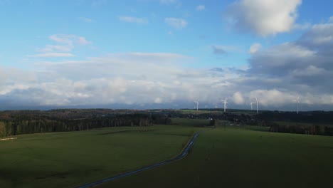 Vista-Aérea-De-Una-Vista-Panorámica-De-Un-Campo-Verde-Con-Un-Camino-Sinuoso,-Bordeado-De-árboles-Y-Turbinas-Eólicas-Bajo-Un-Cielo-Parcialmente-Nublado