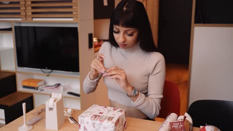 hispanic woman sitting at table decorates and wraps christmas gift, medium shot