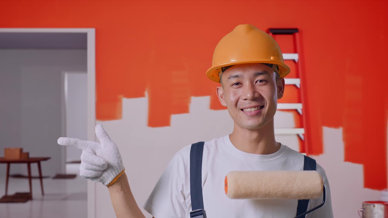 Close Up Of Asian Man Painter Wearing Safety Helmet Smiling And Pointing To Side While Standing With Painting Wall In Room