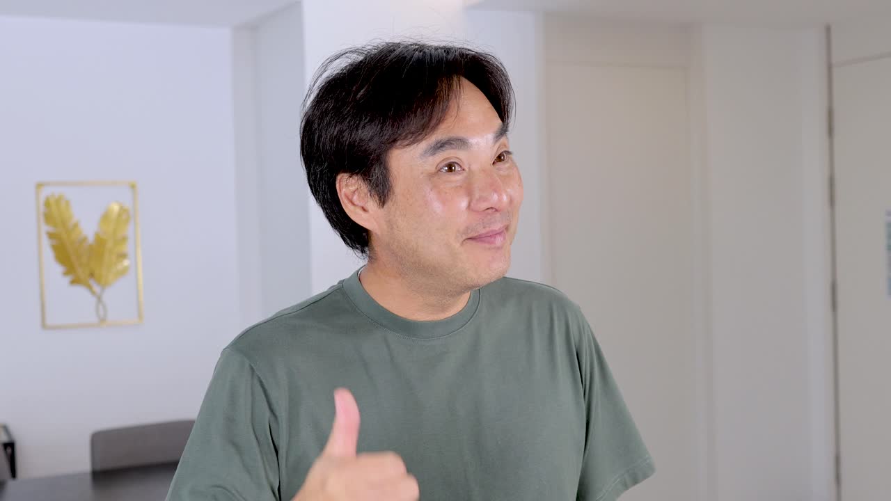 Asian man smiles, makes call me gesture, in bright modern room with soft natural lighting