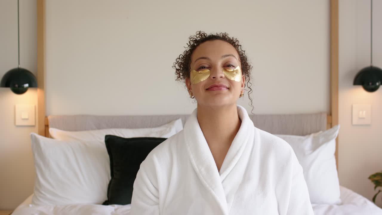 African American woman sitting on bed in white robe wearing gold undereye patches turning, smiling