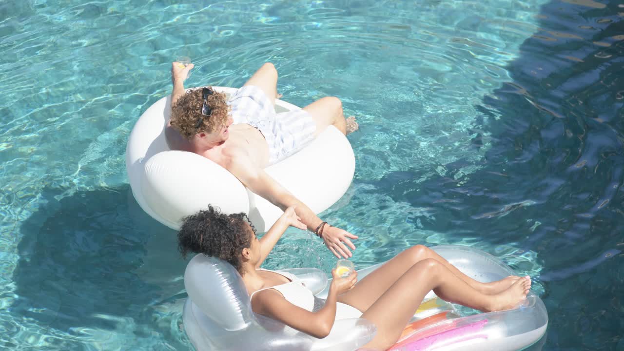 Couple in swimwear floating on pool floats, man shifting reaching for touch while woman sipping cup