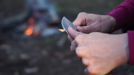 White-female-whittles-a-stick-into-a-spear,-campfire-background,-close-up