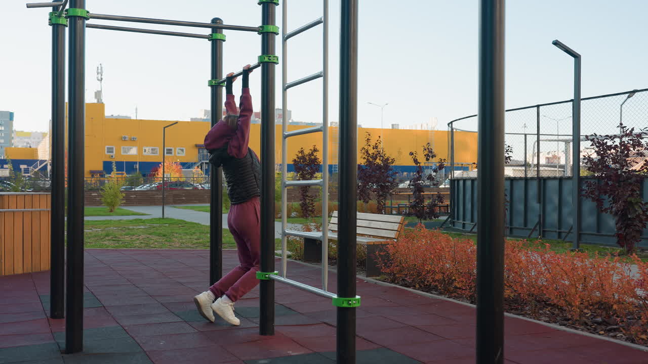Female fit lifestyle advocate placing hands on horizontal bar in urban park outdoor gym wearing burgundy hoodie demonstrating balance control before next pull up with people moving in background