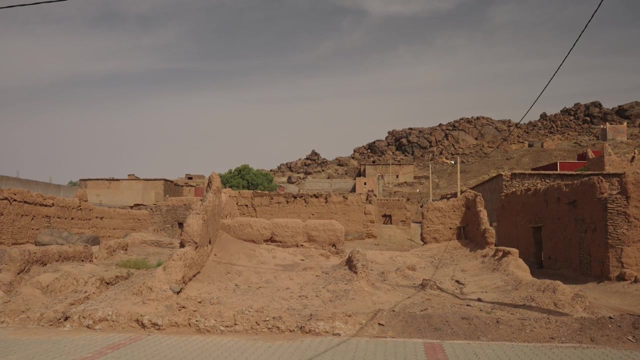 Edifici in mattoni di fango fatiscenti fiancheggiano una strada nel deserto con colline rocciose e cielo grigio nel Marocco rurale.