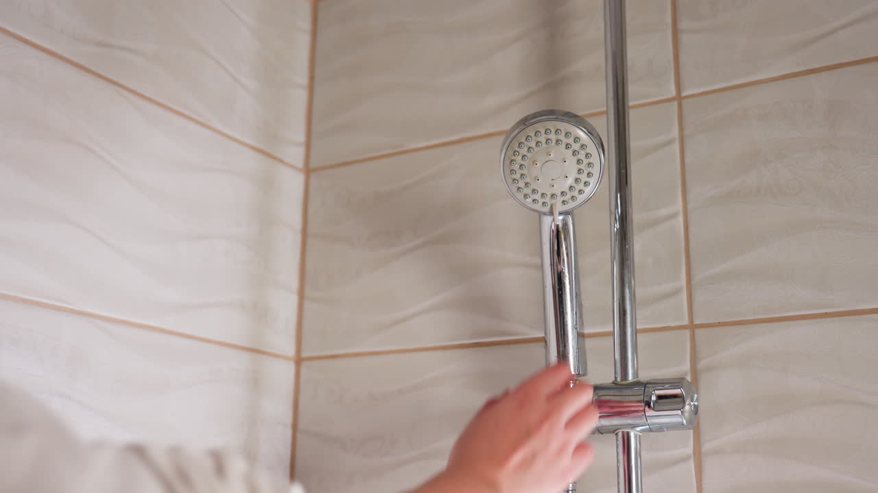 High angle tiled bathroom shows chrome shower on holder as female hand reaches, grips handle, lifts head from mount for washing, light glints on rippled tiles, calm interior, hygiene routine