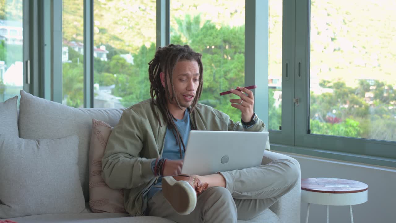 Mid adult man with dreadlocks receiving smartphone call while using laptop, smiling in living room