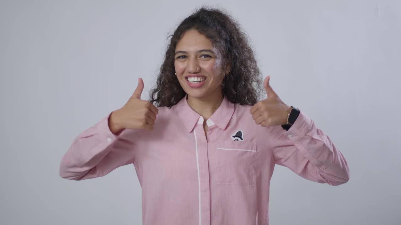 A South Asian woman with curly hair, in a pink shirt, smiles and gives two thumbs up, against a grey background