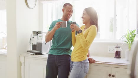 Happy-biracial-couple-standing-in-kitchen,-drinking-coffee-and-laughing