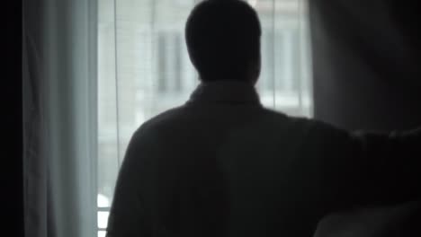 silhouette of a mysterious and stalker-like man opening the curtains of the window of his house to look outside