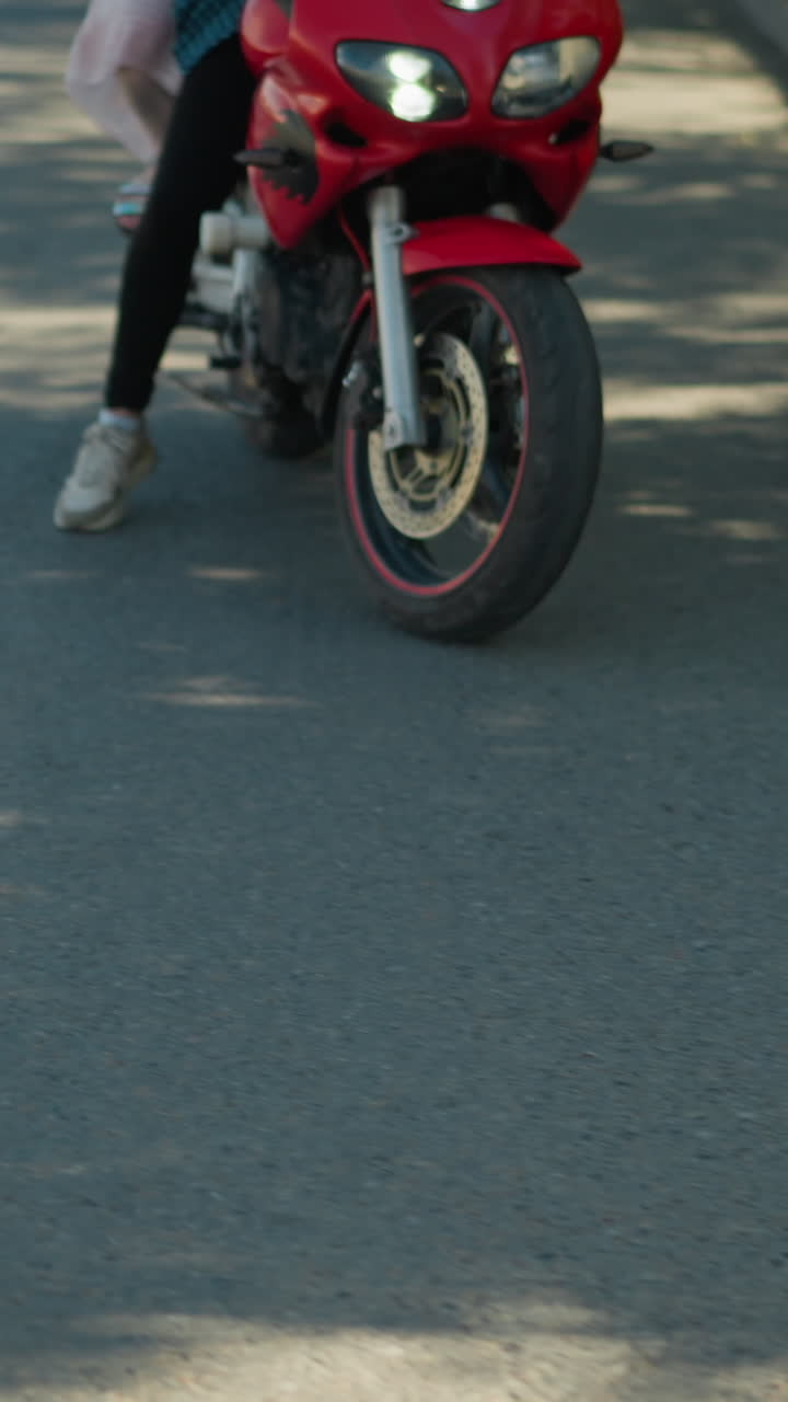 dos mujeres en una bicicleta eléctrica roja navegan por una carretera pavimentada bordeada de árboles verdes exuberantes, un objeto borroso es visible cerca de la cámara, mientras que las sombras y la luz del sol se filtran a través de los árboles